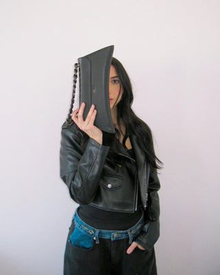 A woman wearing a black leather moto jacket, black top, and blue jeans holds a black leather clutch bag.