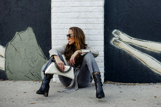 A woman sits on the ground against a painted wall, wearing a light gray coat, gray jeans, and black ankle boots